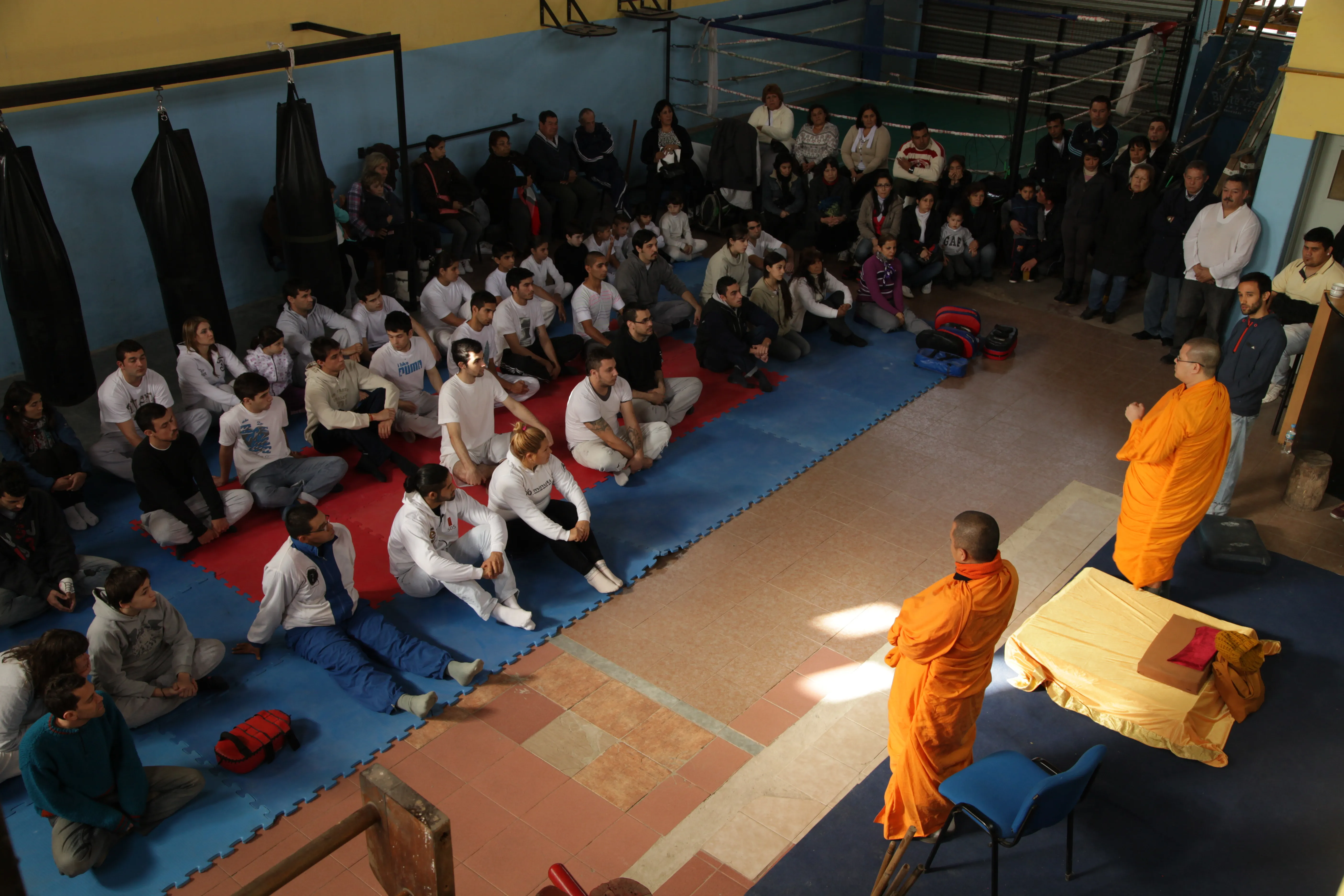 Two monks in front of a gym full of people sitting on mats, seen from above